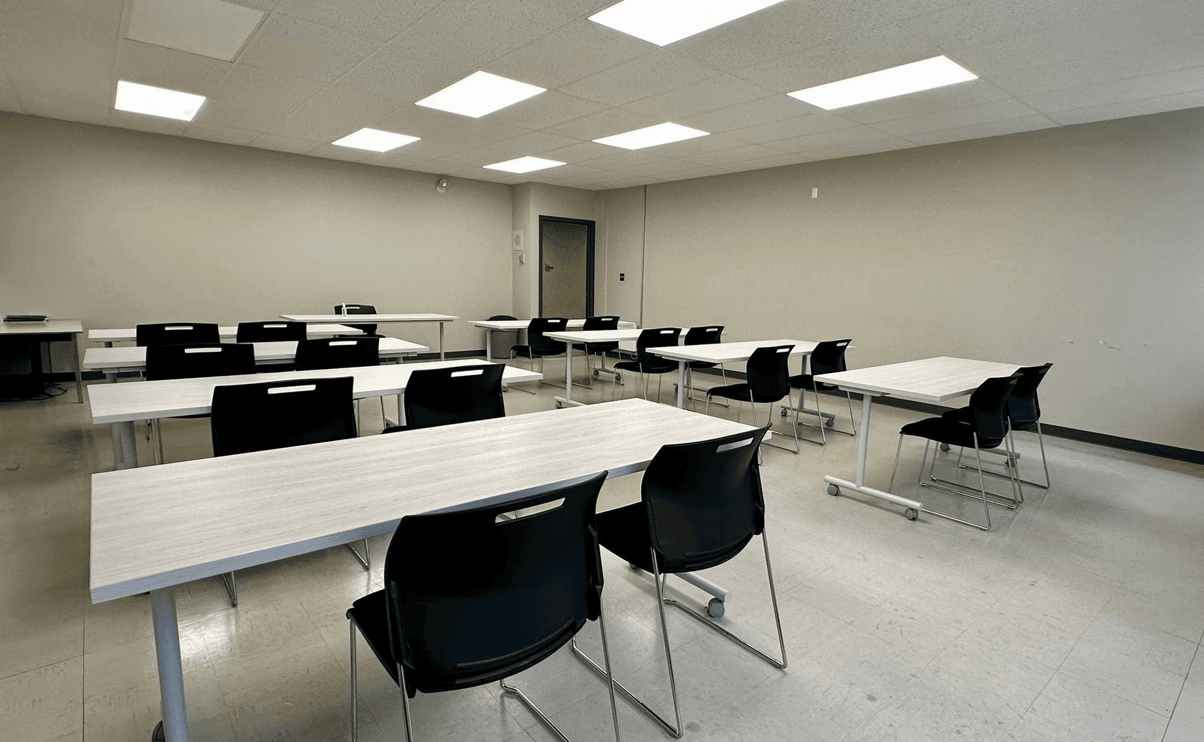 Empty classroom with rows of tables and black chairs under fluorescent lighting.